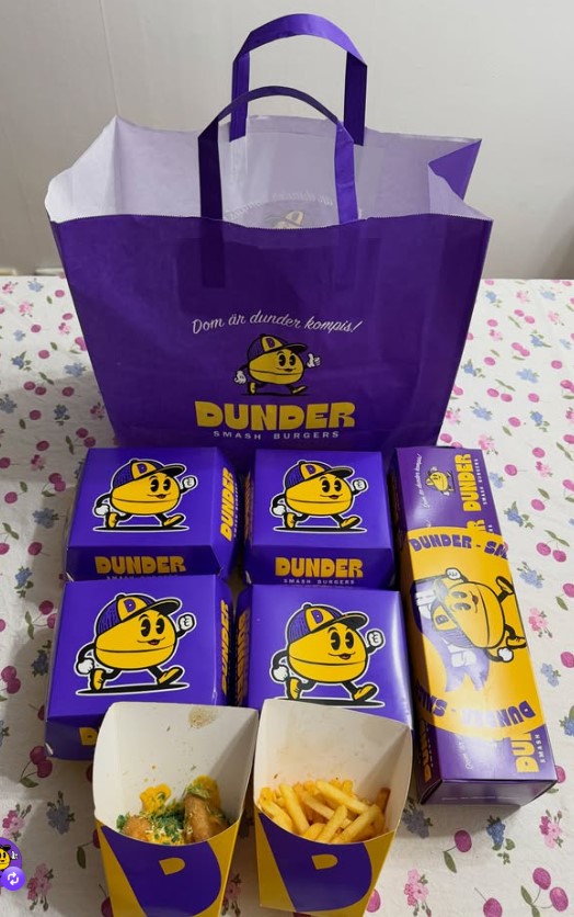 Staplade hamburgerlådor, pommeslådor och påse i lila och gult från Dunder Smash Burgers